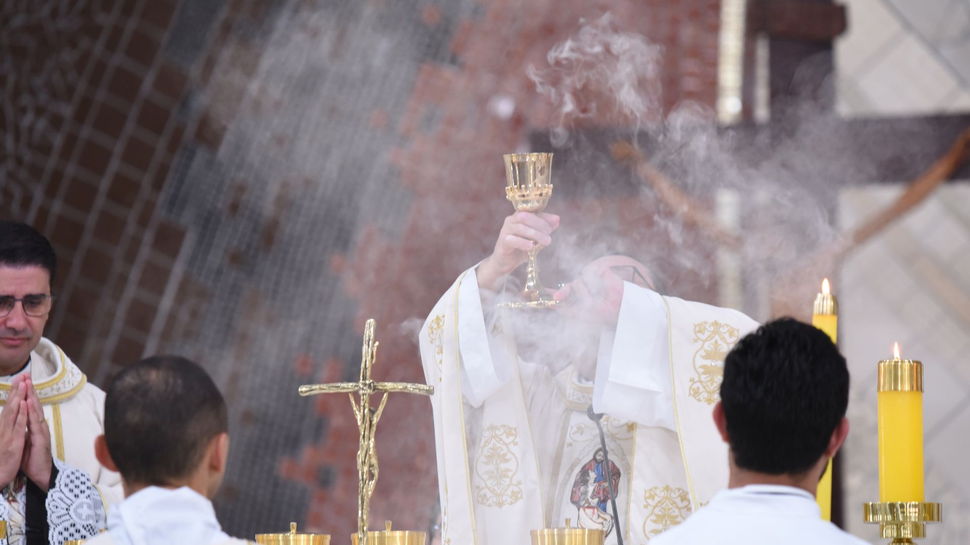 Padre eleva o cálice durante a consagração no santuário do pai das misericórdias, padre ao fundo reza e incenso sobe.