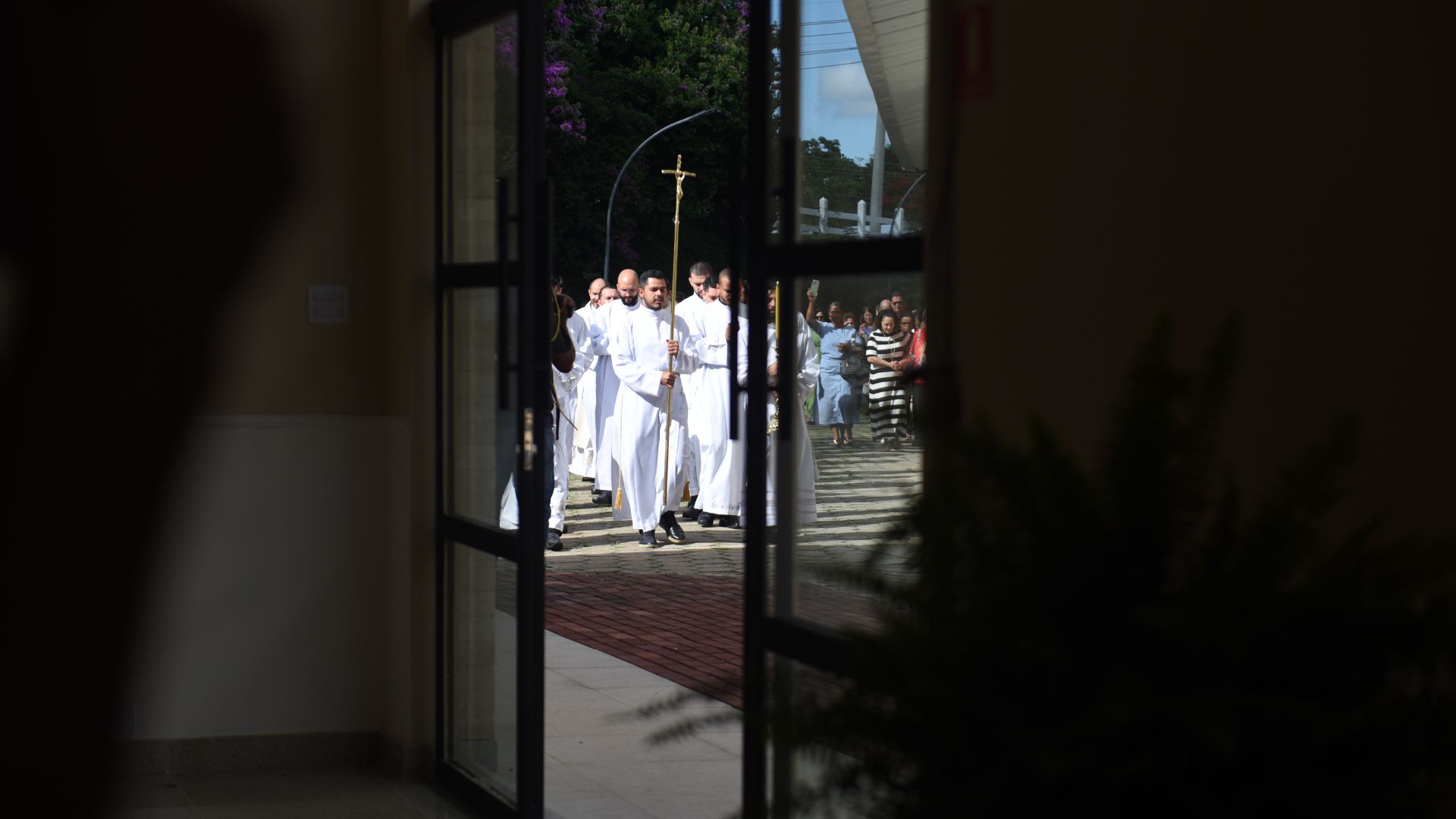 Porta entre-aberta mostra procissão de seminaristas e padres vindo para abertura da nova sala das graças