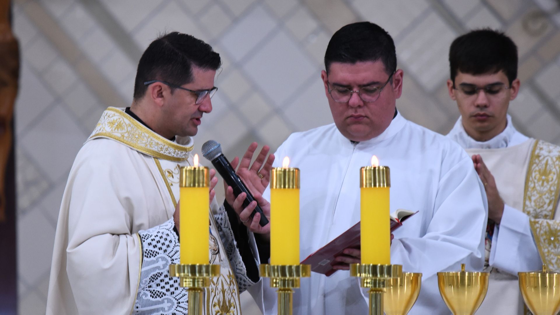 Padre Ricardo faz oração eucaristica durante consagração, velas a frente e seminarista segura livro e padre reza atrás