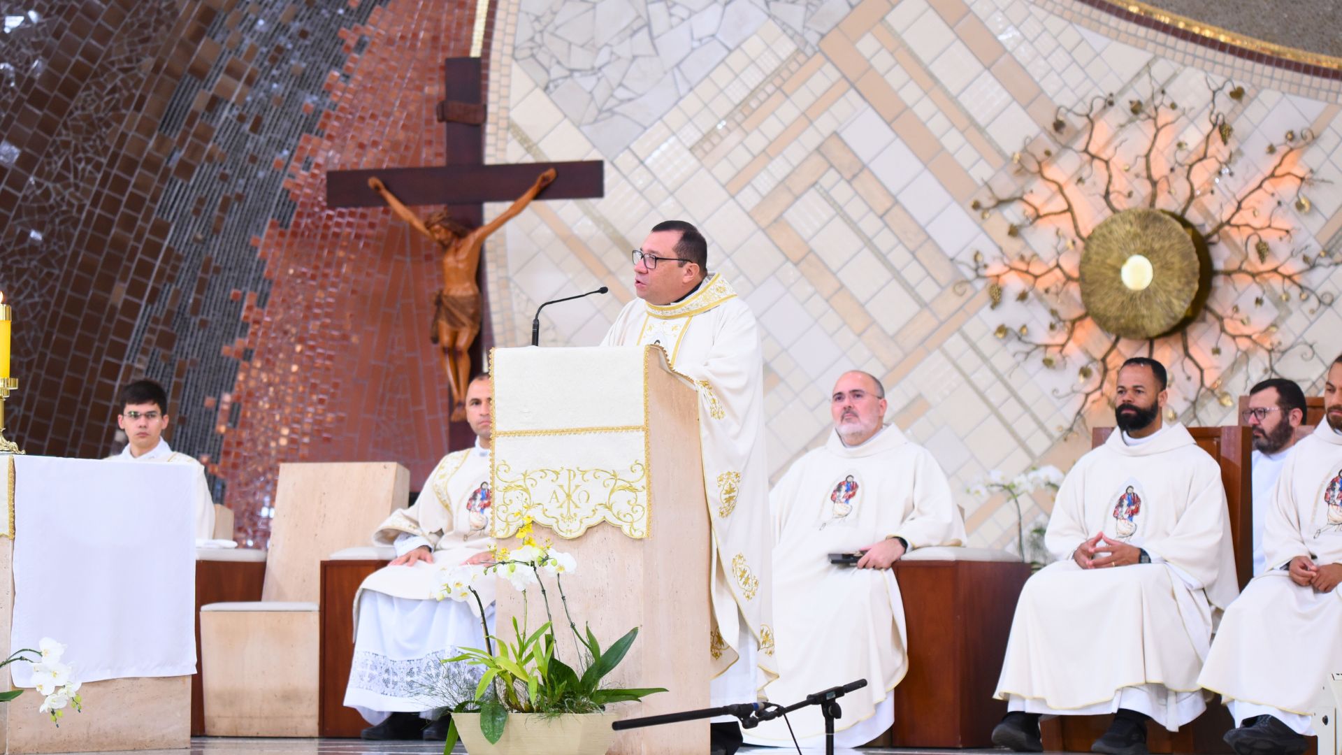 Padre Wagner Ferreira faz homilia no altar central do Santuário do Pai das Misericórdias com sacerdotes sentados ao fundo ouvindo a pregação